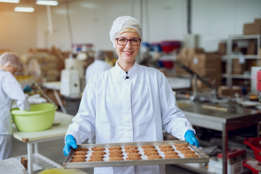 Young happy female worker in sterile cloths holding freshly baked cookies on tinplate inside food production factory.