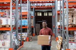 Positive young African-American storeroom worker in glasses walking with boxes over warehouse and carrying order to customer