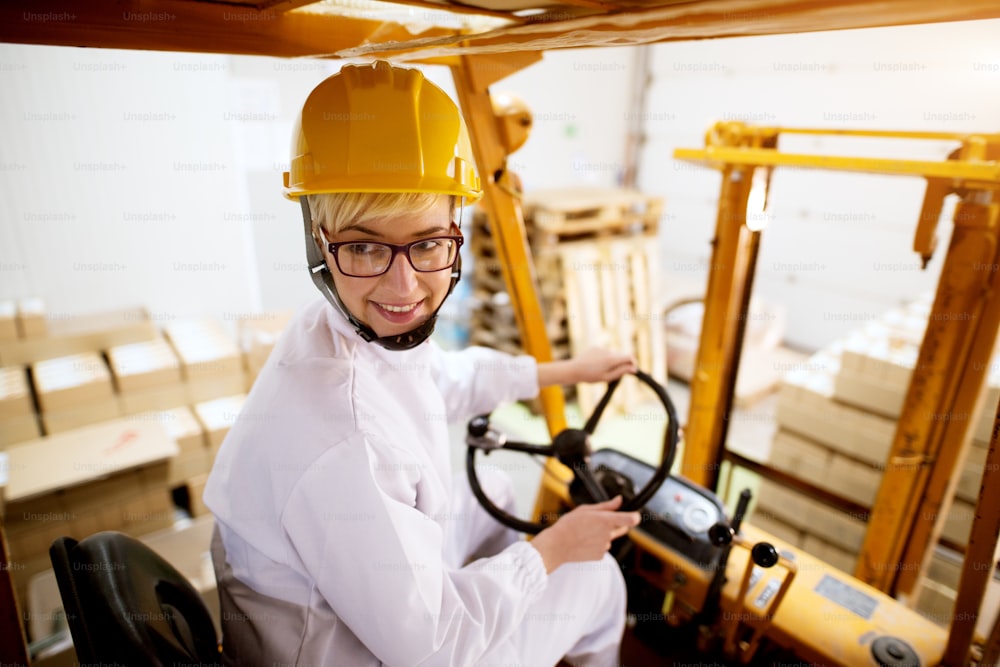Young beautiful happy female worker is driving fork truck in reverse while carrying pallet with a stack of cardboard boxes.