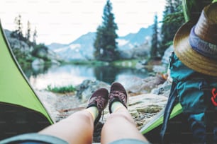 Young woman traveler sitting inside her camping tent with with backpack and enjoying stunning beautiful morning in mountain wilderness near the lake. POV view