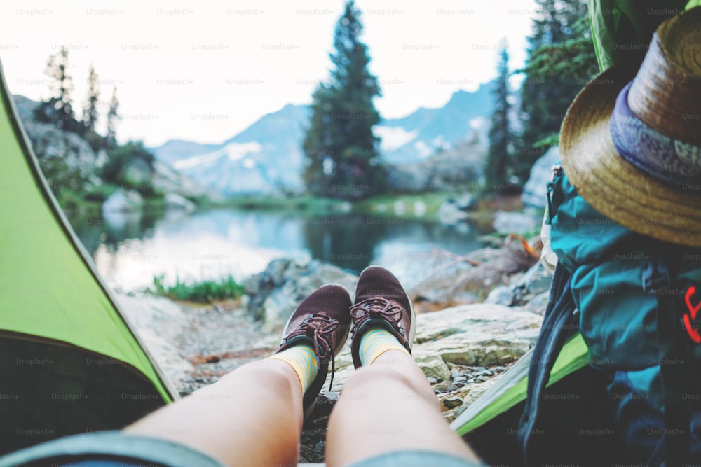 バックパックを背負ってキャンプテントの中に座り、湖の近くの山の荒野で見事な美しい朝を楽しんでいる若い女性旅行者。POVビュー