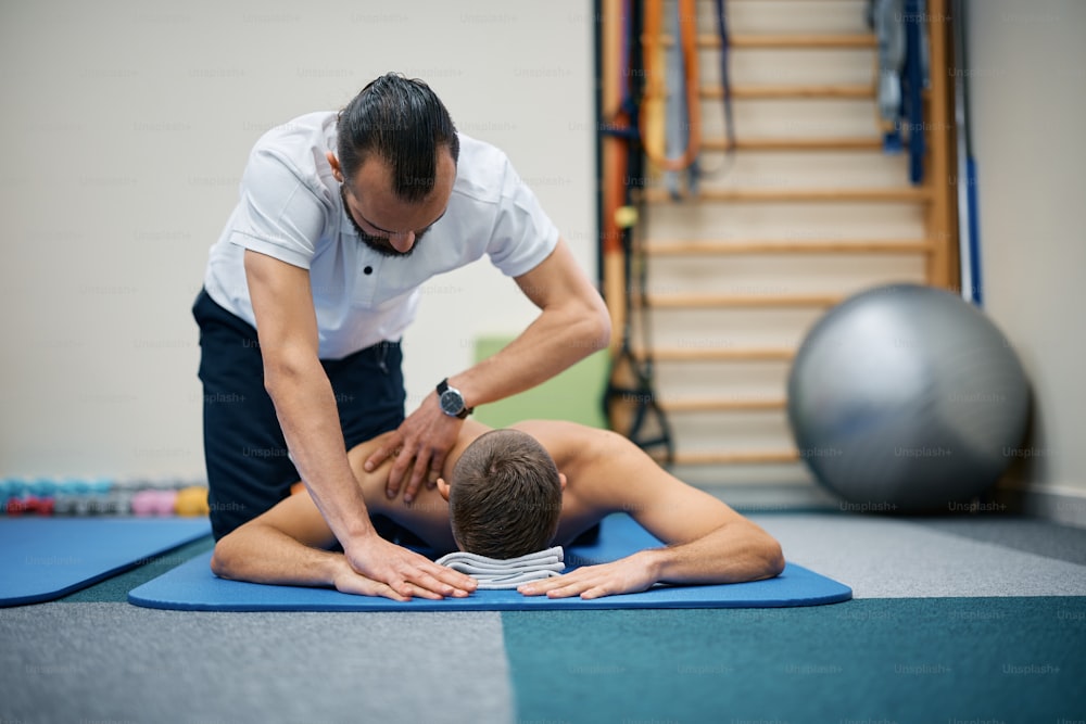 재활 센터에서 어깨를 마사지하는 물리 치료사가 있는 동안 엎드려 누워 있는 운동 남자.