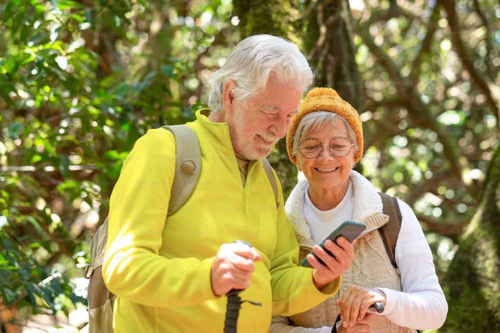 Cheerful caucasian senior couple with backpack and walking sticks hike in the forest while using mobile phone, elderly couple enjoying healthy lifestyle and retirement