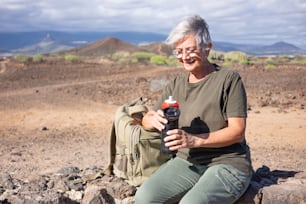 Smiling senior woman on outdoor hike sitting on a stone wall holding her water bottle. Active mature woman sitting near her backpack enjoying freedom and sunny day