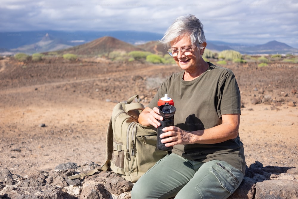 屋外ハイキングで石垣に座り、水筒を持って微笑む年配の女性。自由と晴れた日を楽しんでいる彼女のバックパックの近くに座っているアクティブな成熟した女性