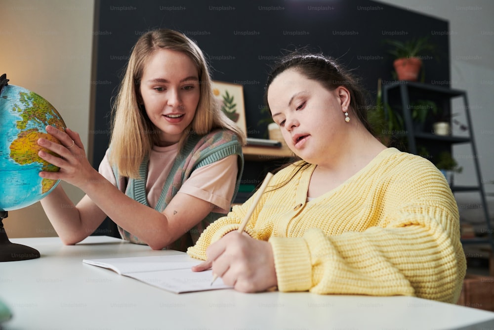 Giovane insegnante che si siede al tavolo accanto al suo studente con sindrome di Down che aiuta con il compito durante la lezione di geografia