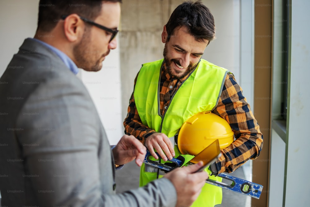 Travailleur de la construction souriant debout avec le superviseur et regardant des plans sur une tablette tout en se tenant dans le bâtiment dans le processus de construction.