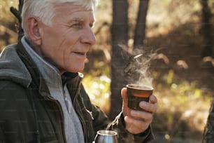 Need some drink. Elderly positive tourist holding cup with hot tea while sitting on the ground in pinewood and looking away. Stock photo