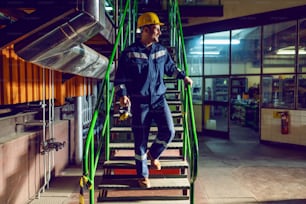 De cuerpo entero de alegre trabajador caucásico vestido con traje de trabajo azul y con casco protector en la cabeza bajando las escaleras. Interior de la planta de energía.