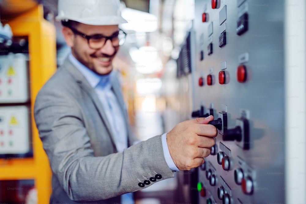 Smiling handsome caucasian supervisor in gray suit and with helmet on head turning switch on. Selective focus on hand. Power plant interior.