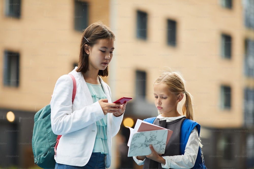 Asian schoolgirl using mobile phone with her younger sister looking at her books while they standing outdoors