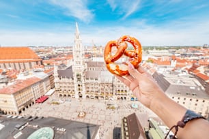 A mão de um turista segura um tradicional lanche alemão Pretzel contra o pano de fundo de uma vista aérea da cidade de Munique a partir do deck de observação
