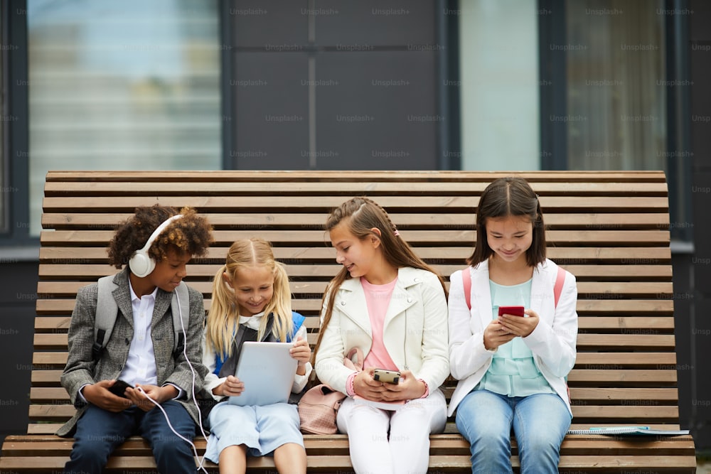 Grupo de crianças em idade escolar sentadas no banco ao ar livre e usando tablet pc e telefones celulares que se comunicam on-line