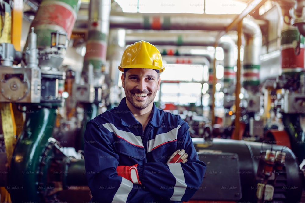 Sorridente trabalhador caucasiano da usina de energia vestido com roupas de trabalho e com capacete na cabeça em pé com os braços cruzados.