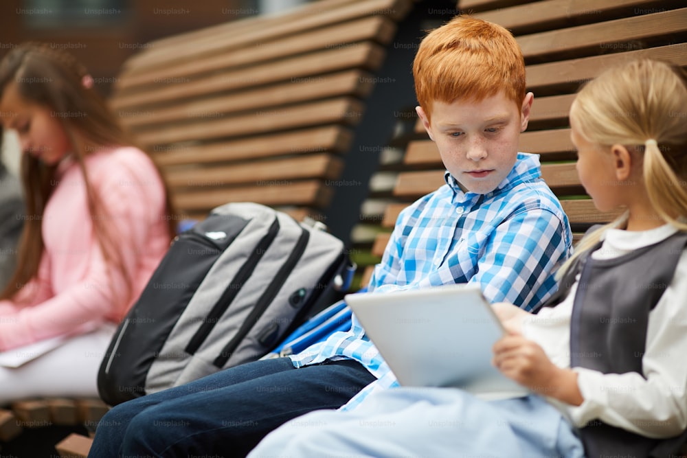 Schoolgirl with tablet pc showing something to the red haired schoolboy while they sitting together on the bench outdoors