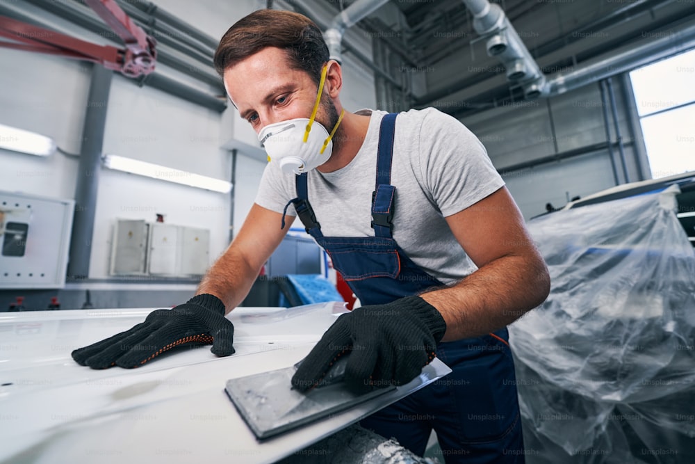 Light vehicle technician removing white paint from car component while working with hand sander