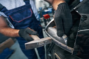 Trabajador que sostiene la pieza de repuesto en el automóvil con una mano mientras lo repara con un martillo en la otra