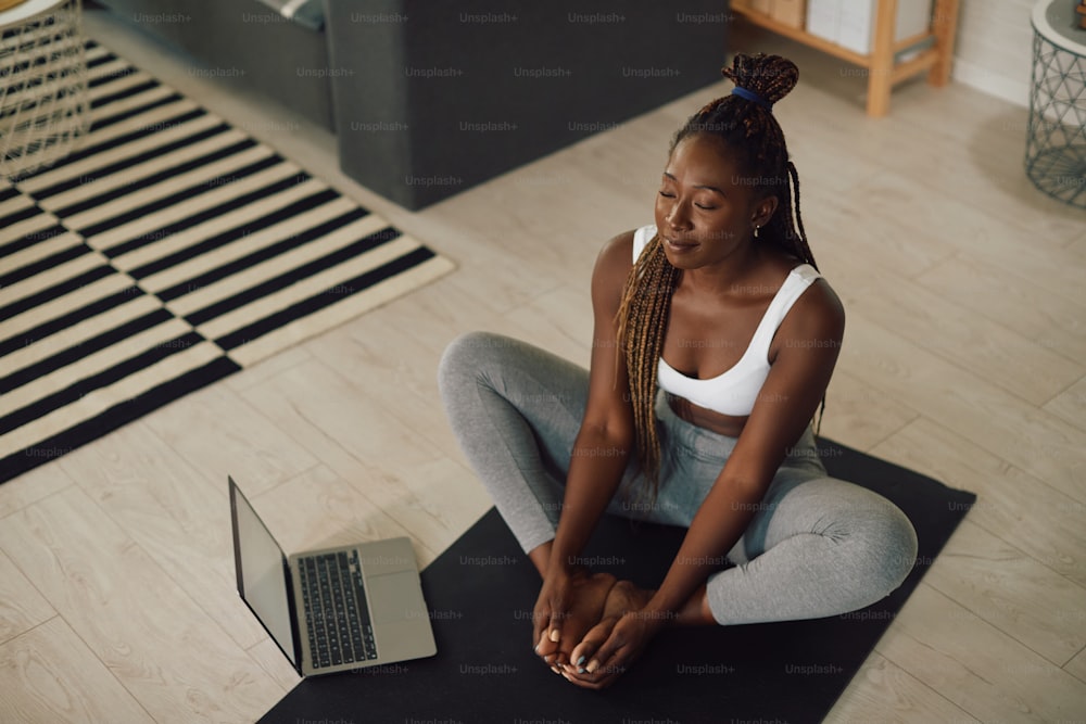 Afroamerikanische athletische Frau, die einen Laptop benutzt, während sie Yoga praktiziert und im Wohnzimmer meditiert.