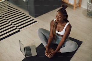 Afroamerikanische athletische Frau, die einen Laptop benutzt, während sie Yoga praktiziert und im Wohnzimmer meditiert.