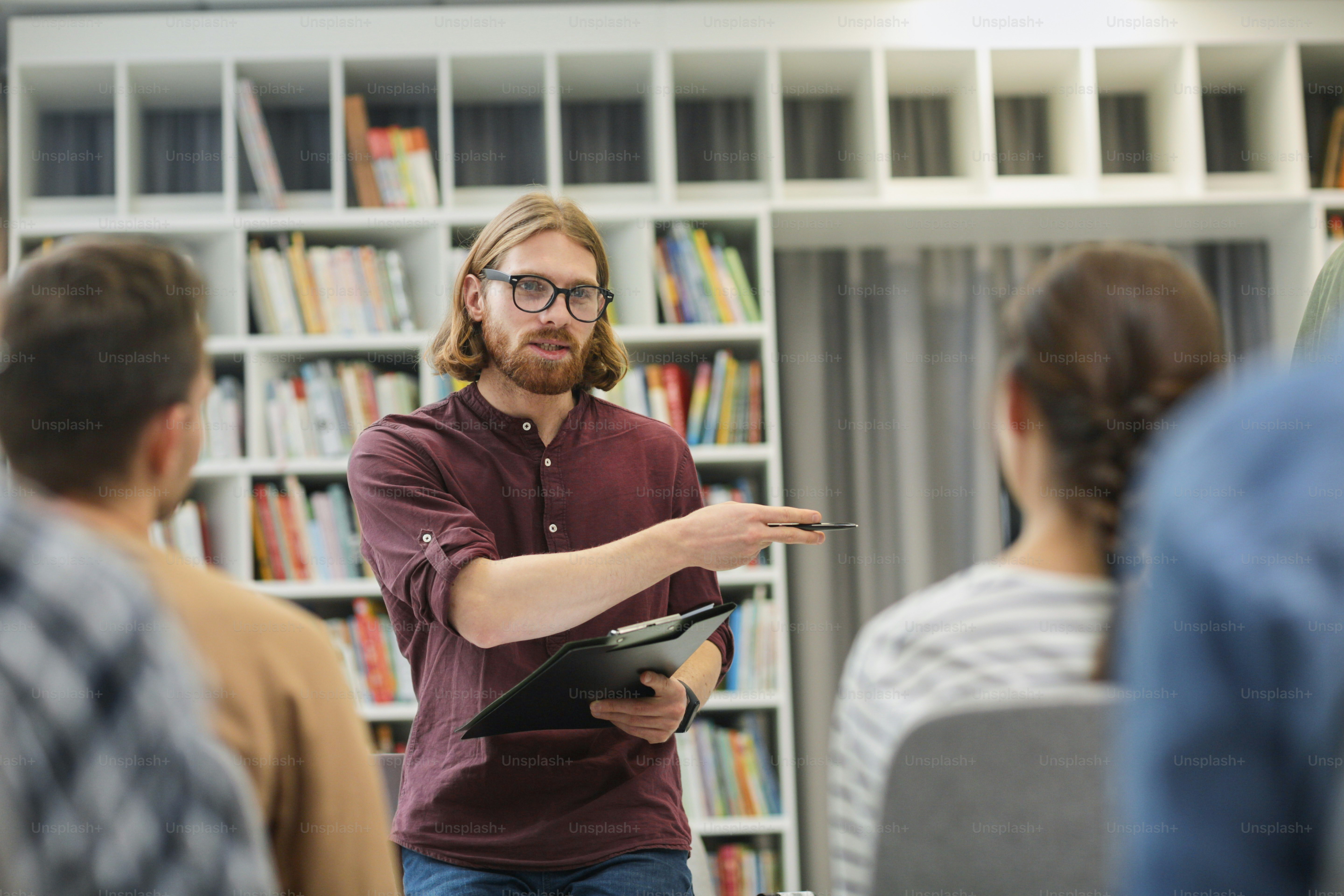 Joven profesor sosteniendo un informe y explicando el nuevo material a los estudiantes durante una lección en la biblioteca