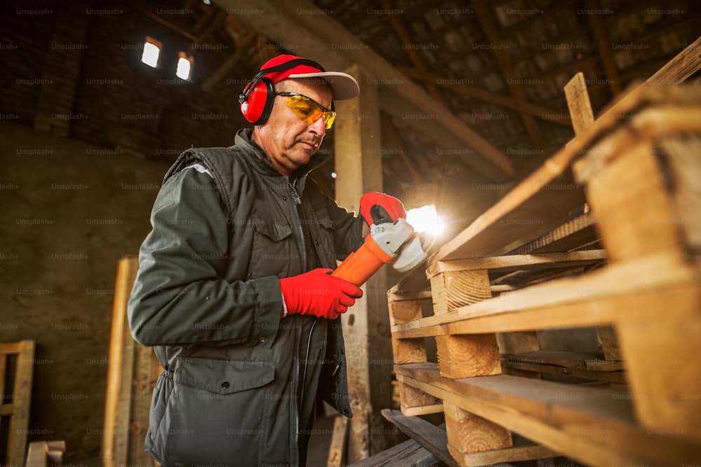 Moderner professioneller Industriearbeiter in Uniform mit Schutz auf der Holzpalette mit der elektrischen Schleifmaschine.