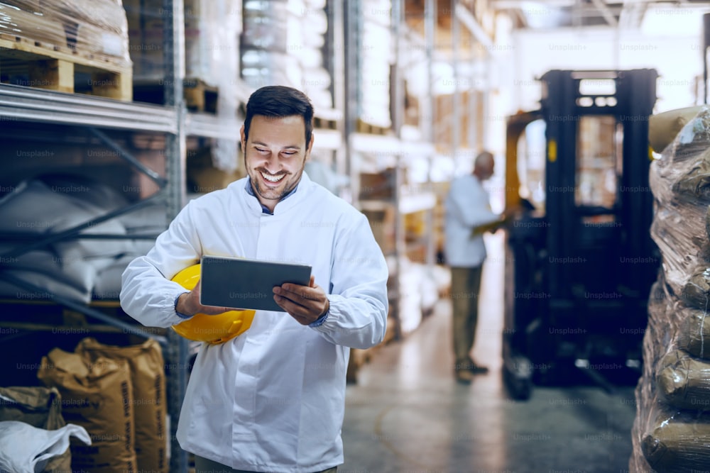 Smiling young Caucasian blue collar worker in white uniform and with helmet under armpit using tablet for controling delivery. In background worker standing next to forklift. Warehouse interior.