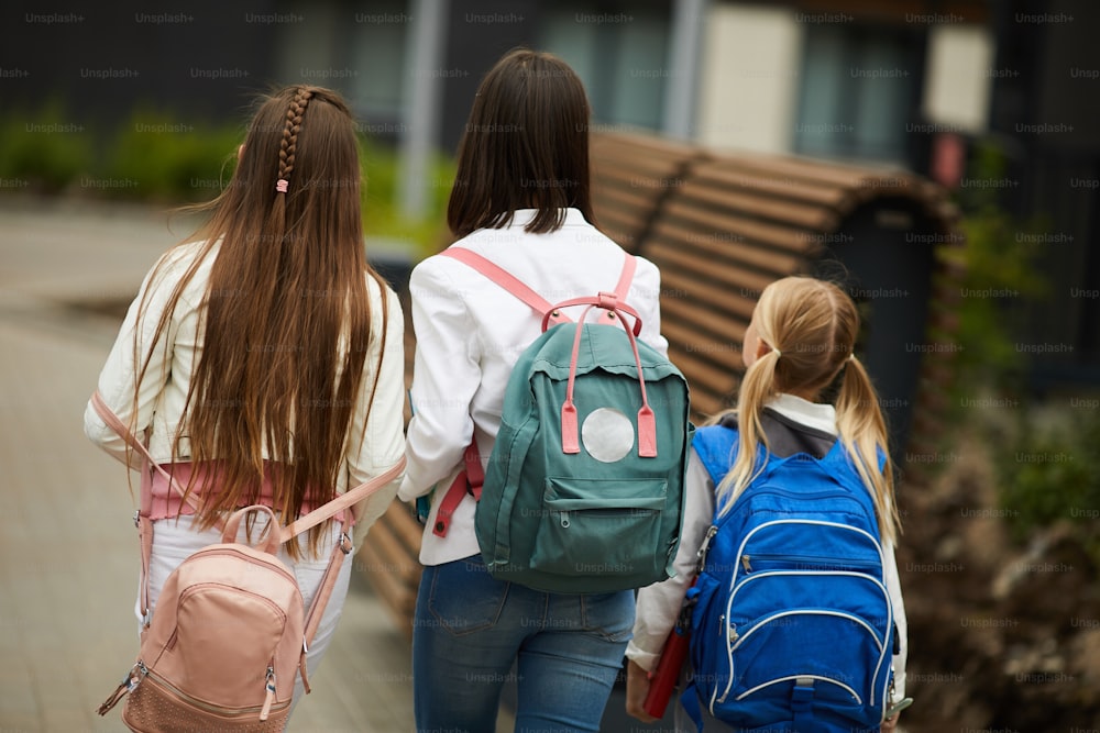 Rear view of schoolgirls with backpacks behind their backs walking together along the street after school outdoors