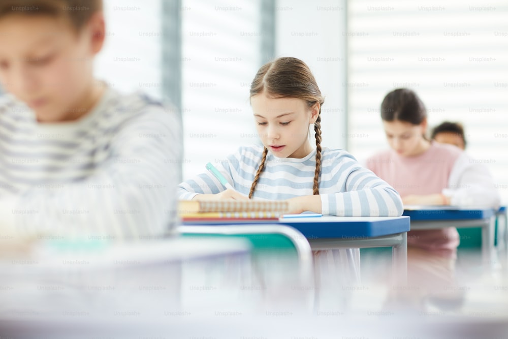 Giovane ragazza caucasica con i capelli castani che indossa una camicia a righe seduta al banco di scuola in un moderno bagno che scrive qualcosa nel suo taccuino, spazio di copia