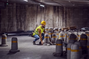 조끼를 쓴 젊은 수염 노동자는 머리에 헬멧을 쓰고 건설 과정에서 터널에 웅크리고 있는 동안 무거운 콘크리트 기둥을 들어 올리려고 합니다.