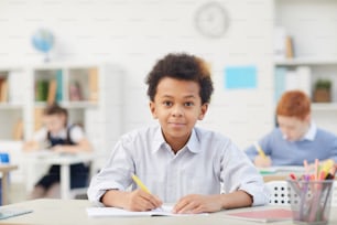Retrato do menino africano sentado na mesa e olhando para a câmera durante a aula na escola