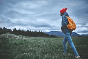 Vue arrière d’une jeune randonneuse faisant de la randonnée dans de belles montagnes. Randonneuse en vêtements imperméables, chapeau rouge et avec un petit sac à dos orange sur fond de pins verts