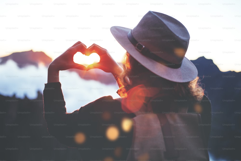 Young traveler woman with backpack and hat standing on edge of cliff at sunset and making by hands in shape of love heart.