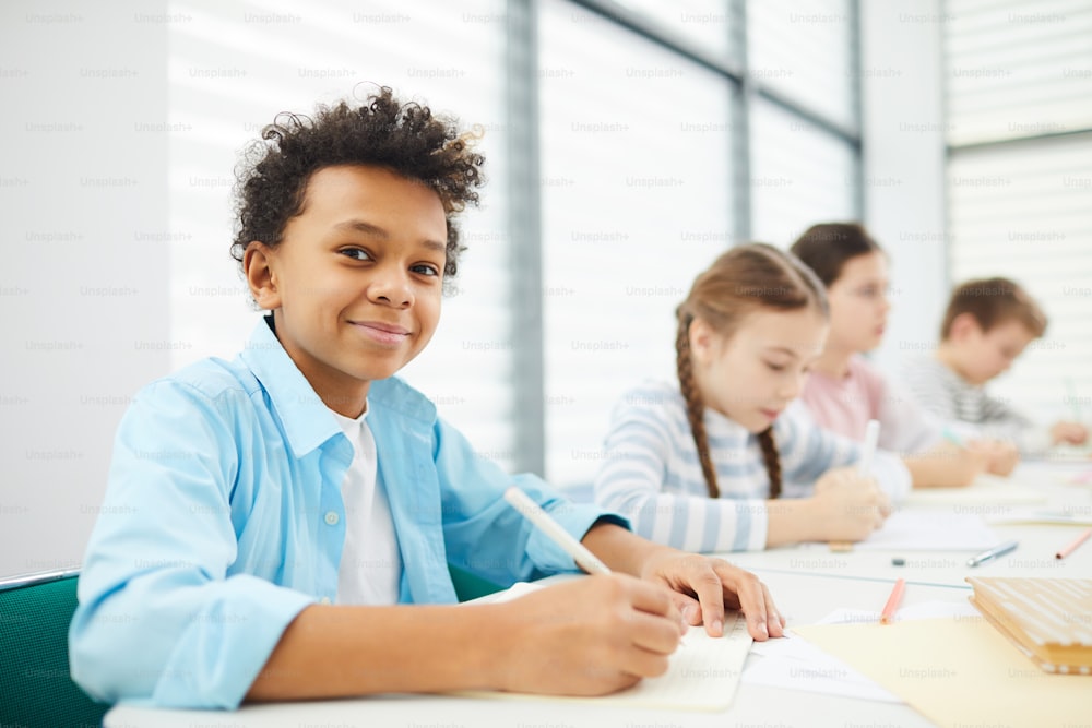Portrait d’un garçon afro-américain joyeux assis au bureau de l’école avec ses camarades de classe regardant la caméra souriante, espace de copie