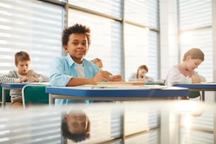 Horizontal shot of young middle school student sitting at desk in modern classroom looking at camera while doing lesson exercise, copy space