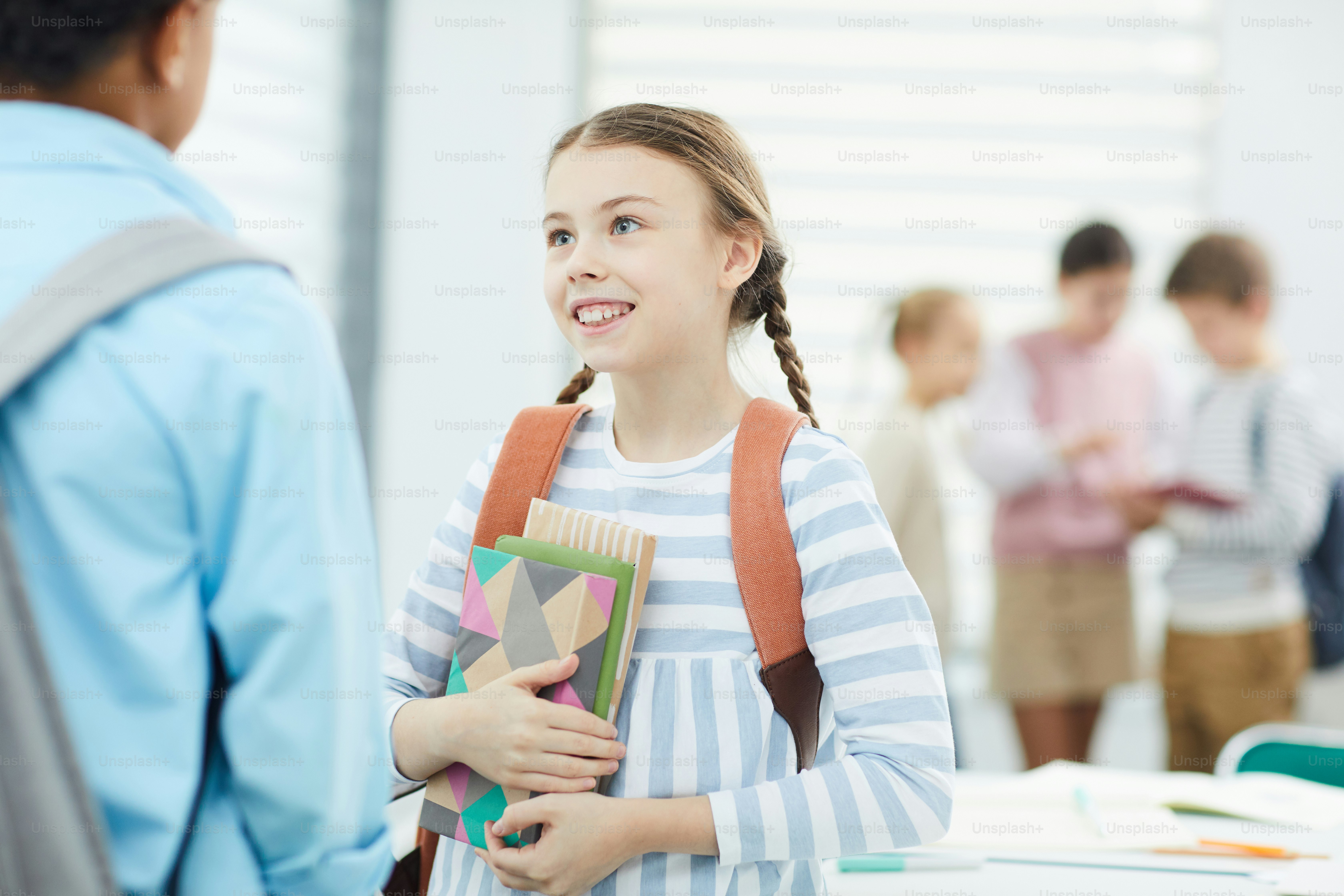 Horizontales mittleres Porträt eines fröhlichen elfjährigen Mädchens mit Rucksack, das in der Pause mit ihrer Schulfreundin plaudert, Kopierraum
