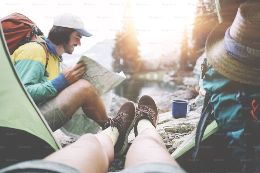 Young couple camping together in stunning mountain wilderness near the lake. POV from the tent. Woman sitting inside, smiling man wearing backpack and searching for trails on the map