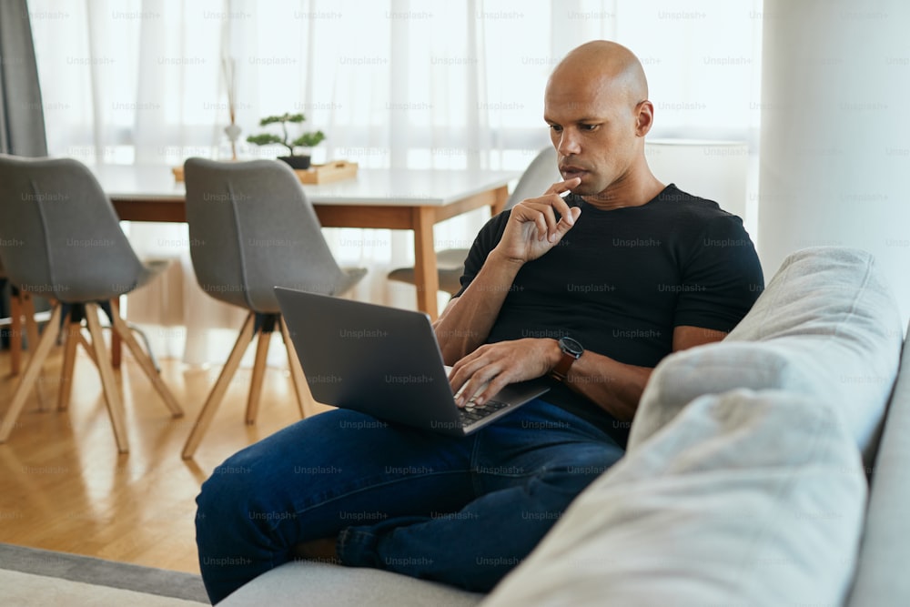 Young African American man thinking while reading problematic e-mail on laptop at home.