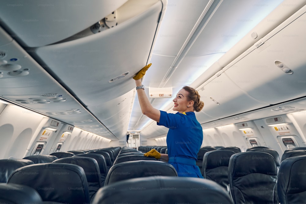 Vista laterale di un assistente di volo carino gioioso in uniforme che chiude il cestino di stivaggio nella cabina dell'aeromobile