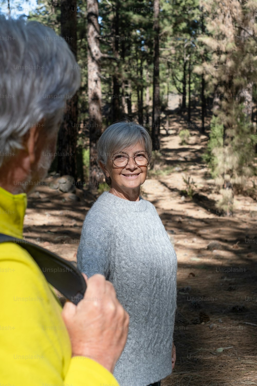 Attractive senior woman looking at his husband walking in the forest holding his hand. Caucasian happy active elderly couple enjoying vacation freedom and nature hiking in footpath
