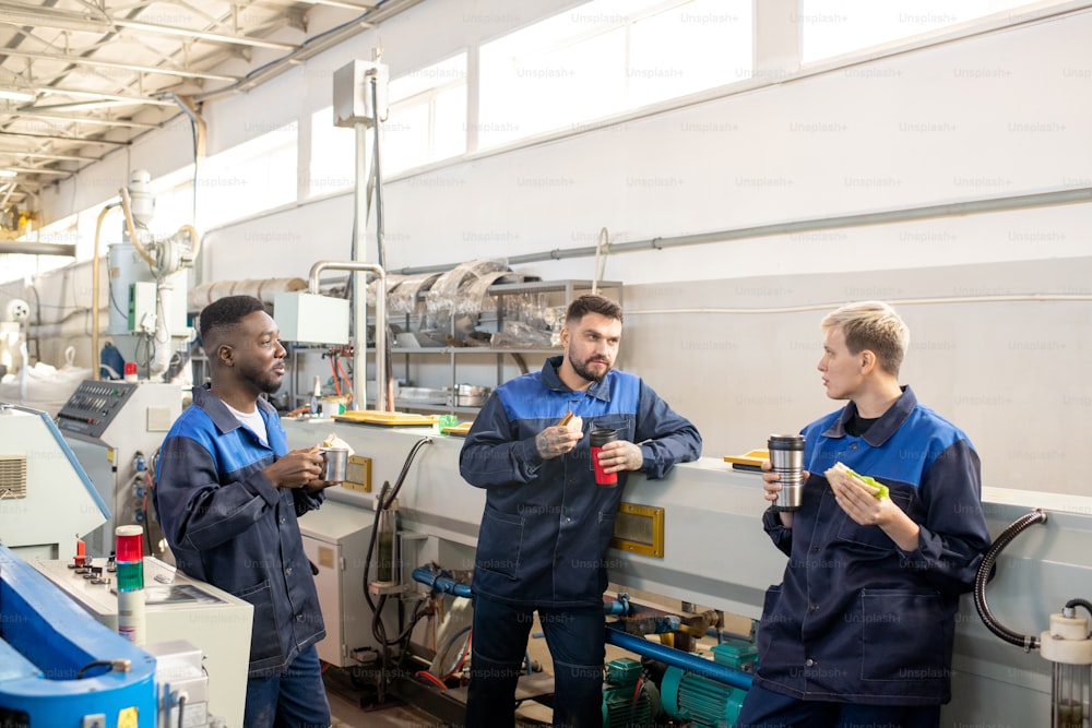 Three young intercultural workers in overalls standing against industrial machine in large workshop, discussing working points and having lunch