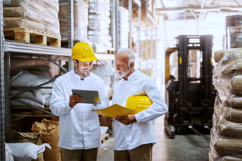 Smiling employees in white uniforms and yellow helmets on heads standing in warehouse and comparing data from tablet. Senior worker holding tablet while younger one holding yellow folder.