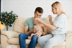 Young father holding baby while mother is feeding him with a bottle at home.