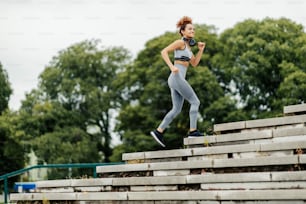 A happy, fit female jogger with headphones around her neck is jogging on the stairs outdoors. An urban jogger practicing outdoors