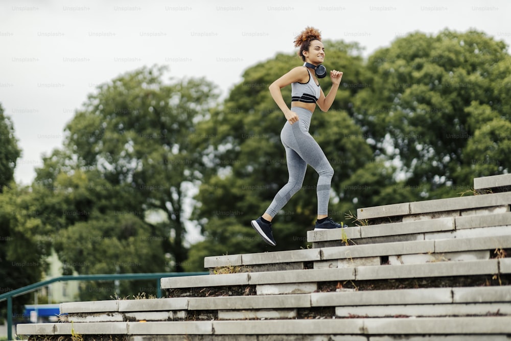 A happy, fit female jogger with headphones around her neck is jogging on the stairs outdoors. An urban jogger practicing outdoors