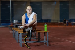 Disable young man in activewear sitting on bench at stadium and using smartphone after training