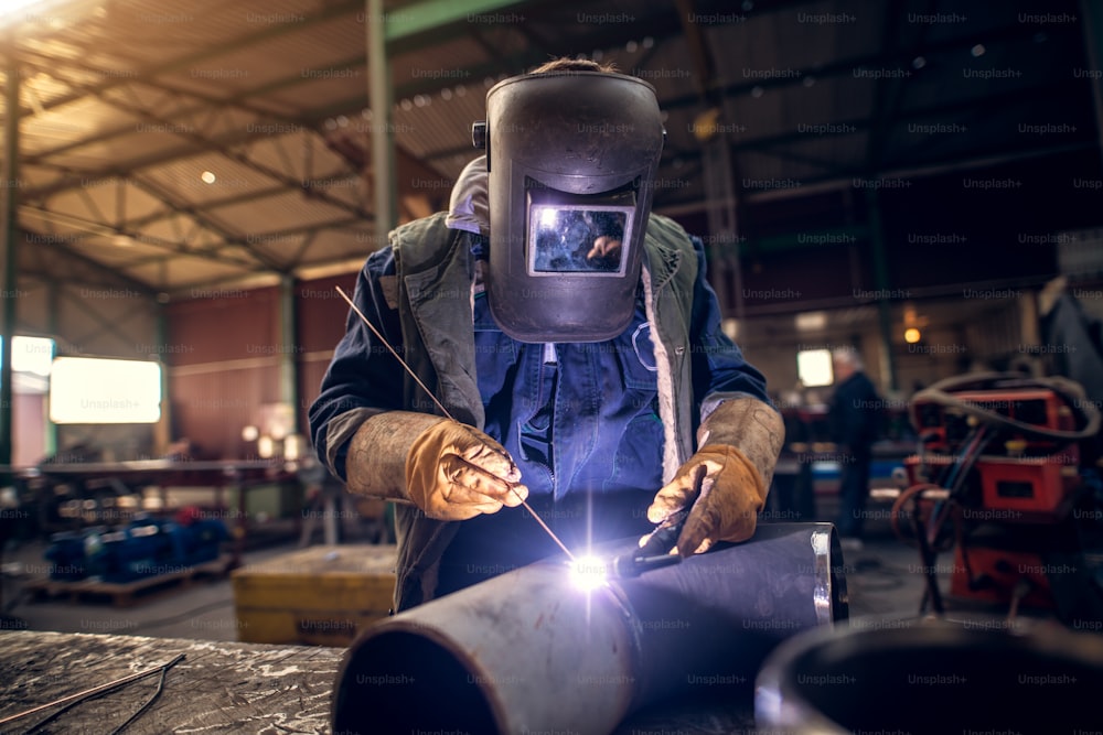 制服を着たプロのマスク保護溶接工の男性が、他の数人の労働者の前で工業用生地のワークショップのテーブルで金属彫刻に取り組んでいるポートレートビューを接写します。