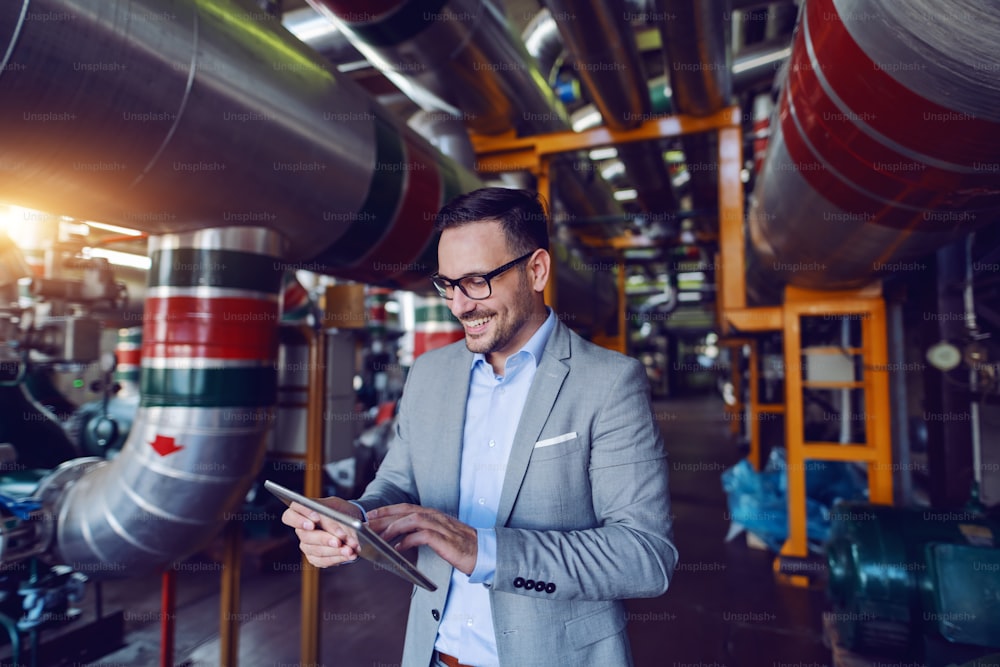 Handsome caucasian manager in gray suit and with eyeglasses using tablet while standing in energy plant.