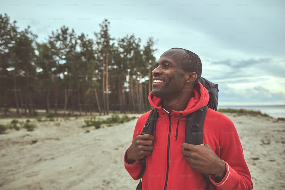 Enjoying nature. Waist up portrait of man standing on the shore and looking at the forest. He holding his backpack with both hand. Copy space on the left side