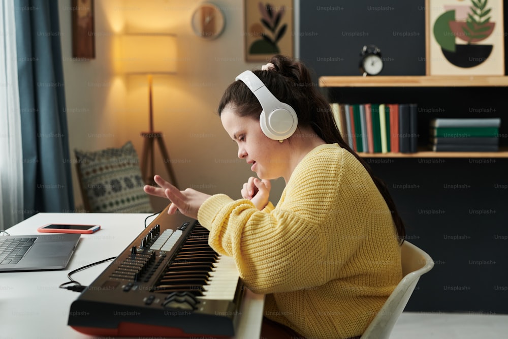 Modern young Caucasian woman with Down syndrome spending time at home composing song using electronic keyboard