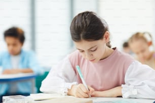 Twelve-year-old schoolgirl with dark brown hair sitting at school desk writing essay, horizontal medium close up portrait